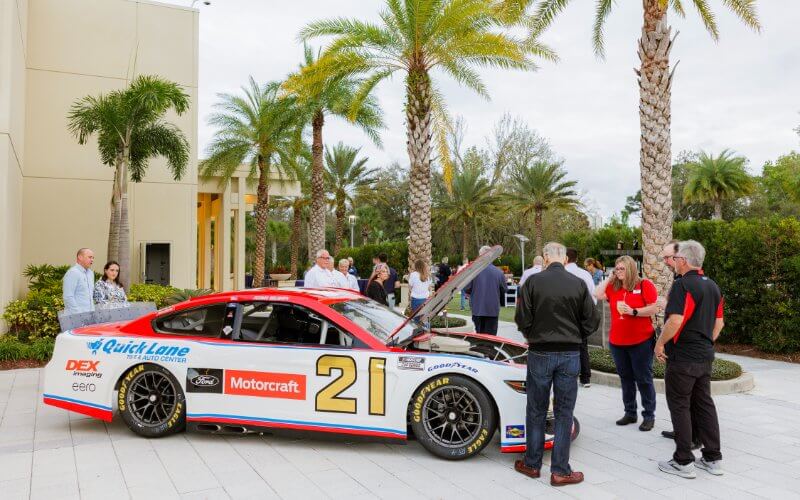 A red and white race car with the number 21 is parked outside, with its hood raised. People are gathered around, engaging in conversation against a backdrop of palm trees and a building.