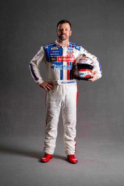 Josh Berry in his racing suit on a grey background holding his helmet in one hand